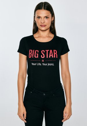 Hosszú, sötét hajú nő fekete "BIG STAR Your Life. Your Jeans." feliratú pólóban és fekete nadrágban, sima világos háttér előtt áll.