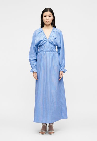 Γυναίκα στέκεται φορώντας μακρύ μπλε ριγέ φόρεμα με ραμμένη μπούστο και μέση, μακριά φουσκωτά μανίκια και λευκά σανδάλια με ανοιχτά δάχτυλα.