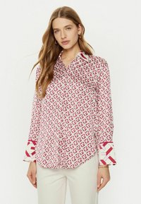 Camicia bianca con un motivo geometrico rosso, colletto a punta e polsini a contrasto con un design simile. Tessuto morbido, stile con bottoni.