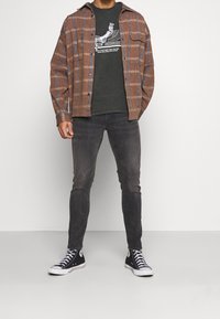 Hnědá kostkovaná košile s modrými pruhy, šedé tričko s grafikou, tmavé džíny a černé vysoké tenisky. Neformální oblečení s vrstvenými texturami.