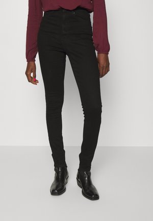 Person iført sorte højtaljede skinny jeans, sorte ankelstøvler og en vinrød top med lange ærmer, stående mod en ensfarvet baggrund.