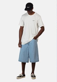 Camiseta blanca de manga corta con un pequeño gráfico, pantalones cortos de mezclilla azul claro y gorra negra con bordado blanco; zapatillas negras con suelas beige.