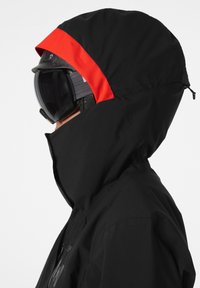 Black ski jacket with a red accent on the hood. Zippered front and high collar. Includes snow goggles on the wearer. Matte finish texture.