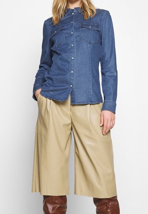 Spijkershirt in medium blauw, met knoopsluiting, puntige kraag en borstzakken. Gecombineerd met beige broek met wijde pijpen en bruine laarzen.