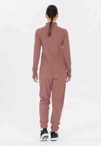 Vrouw met ingevlochten haar, gekleed in een mauve lange mouwen top en broek, zwarte en witte sneakers, staand met haar rug naar de camera tegen een plain witte achtergrond.