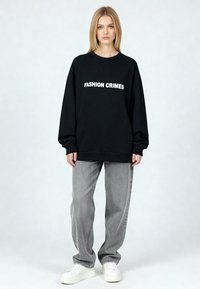 Sweat-shirt noir avec texte blanc "FASHION CRIMES", coupe décontractée. Associé à un jean gris ample et des baskets blanches ; texture lisse.