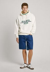 Felpa bianca con testo verde, pantaloni shorts in denim e sneakers bianche, caratterizzata da un design sportivo e una vestibilità rilassata. Dettagli notevoli includono il logo e le cuciture.