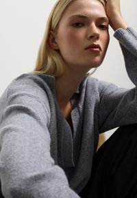 Grauer Strickpullover mit V-Ausschnitt und gerippten Bündchen, im Close-up präsentiert. Weicher Stoff und bequeme Passform, kombiniert mit schwarzen Hosen.