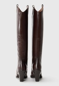 Botas de cuero marrón hasta la rodilla con un diseño elegante, punta afilada y un tacón modesto; cuentan con dos bucles en la parte superior para facilitar su uso.