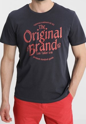 T-shirt en coton bleu foncé avec graphismes imprimés rouges, comprenant le texte "The Original Brand" et un logo. Manches courtes, col rond, coupe décontractée.
