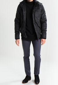 Black padded jacket with a high collar, front pockets, worn over a black turtleneck. Paired with slim grey pants and black shoes.