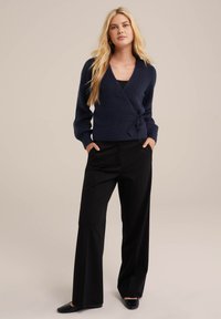 Temno modri pleteni ovitek s dolgimi rokavi in detajlom s povezovanjem, v kombinaciji z črnimi širokimi hlačami in ravnimi črnimi čevlji.