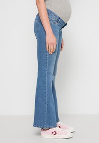 Svetlo modre razširjene nosečniške jeans hlače s srednje visokim pasom, narejene iz jeansa, z žepi in gladko teksturo. Kombinirane z rožnatimi supergami.