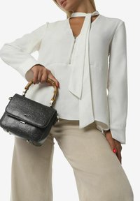 Borsa nera con motivo in rilievo, dotata di un manico in bambù, indossata con una blusa bianca e pantaloni beige. La blusa ha un dettaglio con zip e dei lacci.