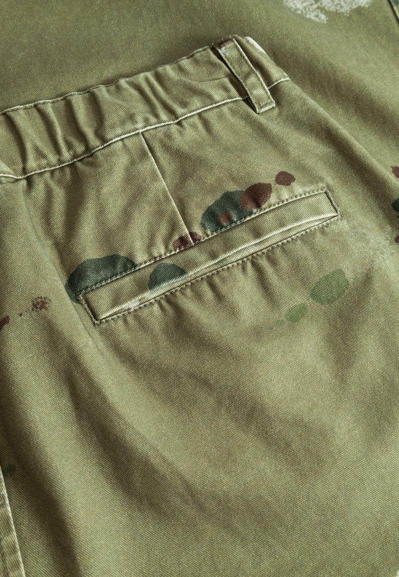 Grøn stof med en baglomme, der har et plisseret design. Flere malerisk lignende pletter i brun og grøn tilføjer mønsterdetaljer.