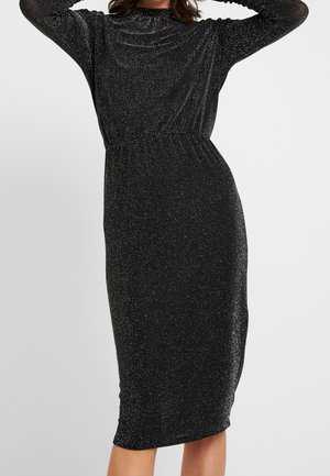 Femme portant une robe midi noire ajustée avec manches longues et tissu scintillant, posant avec les mains derrière la tête sur un fond uni.