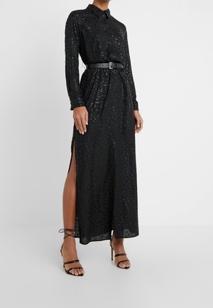 Kvinde iført en lang sort leopard print skjortekjole med sideslid, sort bælte og sorte strop-sandaler med høj hæl.