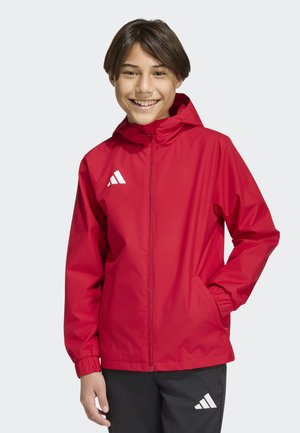 Jeune souriant portant une veste zippée à capuche rouge et un pantalon de sport noir avec un logo blanc, debout avec une main dans la poche.