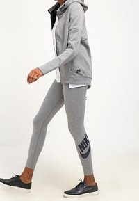 Osoba v šedých legínách Nike s logem na lýtku, odpovídající zipové mikině, bílém tričku a černých teniskách, jde proti jednoduchému pozadí.