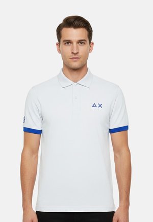 Uomo che indossa una polo bianca con rifiniture blu sulle maniche e logo a triangolo e X blu sul petto sinistro, rivolto verso avanti su sfondo bianco.