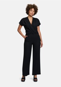 Schwarzer Jumpsuit mit kurzen Ärmeln, tiefem V-Ausschnitt und weitem Bein. Glatter Stoff mit taillierter Passform, der subtile Drapierungsdetails aufweist.