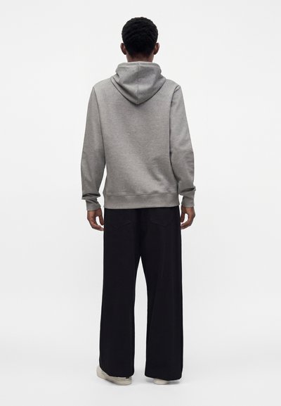 Personne debout de dos, portant un sweat à capuche gris, un pantalon large noir et des chaussures claires sur fond blanc.