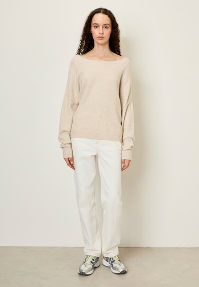 Femme aux cheveux bouclés portant un pull beige surdimensionné, un pantalon blanc et des baskets grises, debout contre un mur clair uni.