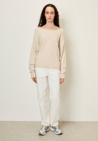 Beige gebreide trui met een brede halslijn, afhangende schouders en lange mouwen, gecombineerd met witte rechtpasvorm jeans en veelkleurige sneakers.