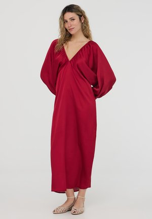 Lungo vestito rosso scuro con scollo a V e maniche a sbuffo. Tessuto liscio, lunghezza maxi e dettagli minimali. Sandali casual completano il look.