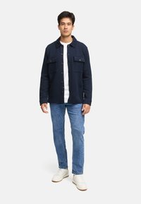 Marineblaue gesteppte Jacke mit Kragen, zwei Brusttaschen und braunen Knöpfen, kombiniert mit hellblauen Jeans und weißen Turnschuhen.