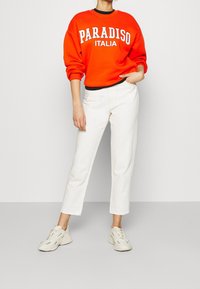 Červená mikina s potiskem "PARADISO ITALIA" v kombinaci s bílými zkrácenými kalhotami. Oblečení doplňují světlé tenisky. Jednoduchý design, neformální styl.