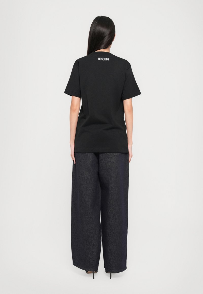 Schwarz Baumwoll T-Shirt mit kurzen Ärmeln und Rundhalsausschnitt, mit einem weißen "MOSCHINO" Logo auf der oberen Rückseite bedruckt. Kombiniert mit weitem, dunklen Denim-Hosen.