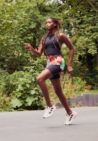 Un débardeur de sport noir et un short à motifs colorés. Les chaussures de course sont blanches avec des rayures noires et des accents roses. Un feuillage vert en arrière-plan.