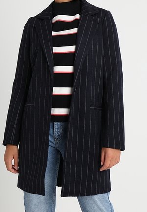 Person trägt einen marineblauen Nadelstreifenmantel über einem schwarz-weiß-roten gestreiften Pullover mit hellblauen Jeans vor einem schlichten Hintergrund.