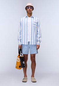 Cămașă cu dungi albastre și albe, mâneci lungi, șorturi albastre deschise, pantofi slip-on bej, pălărie roz de tip bucket și ochelari de soare, ținând două sacoșe.