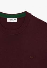 Maglione a collo alto color bordeaux con colletto a coste verde. Presenta il logo Lacoste ricamato in marrone scuro. Realizzato in tessuto lavorato a maglia leggero e testurizzato.