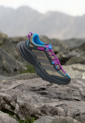 Zapatilla de trail running con detalles en gris, azul y púrpura sobre terreno rocoso y fondo de montaña desenfocado.