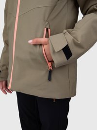 Persoană îmbrăcată cu o jachetă taupe cu fermoar roz, având mâna în buzunarul lateral cu fermoar, pantaloni negri vizibili pe un fundal luminos simplu.
