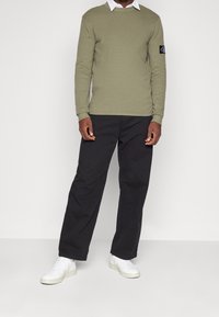 Chemise à manches longues vert olive avec un col rond, associée à un pantalon large noir et des baskets blanches. Présente un patch logo sur la manche.