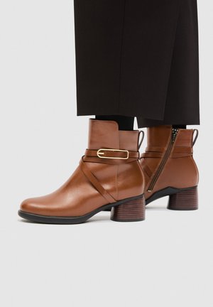 Stivaletti alla caviglia in pelle marrone con cerniera laterale, cinturino con fibbia dorata, tacco basso impilato, indossati con pantaloni neri e calzini neri.