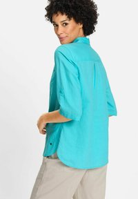 Camicia di lino turchese con colletto, maniche a tre quarti e orlo curvo. Presenta una piega sul retro e un piccolo logo dettagliato sul lato.