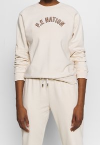 Krämfärgad sweatshirt med ribbad mudd, rund halsringning och broderad "P.E. NATION" i brun tråd. Matchas med ljusa byxor.