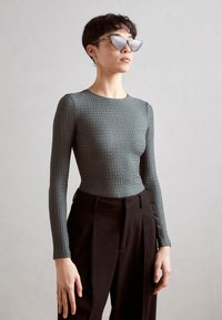 Graues, strukturiertes Langarmoberteil, kombiniert mit schwarzen, hochgeschnittenen Hosen. Das Model trägt Katzenaugen-Sonnenbrillen und einen Creolenohrring.