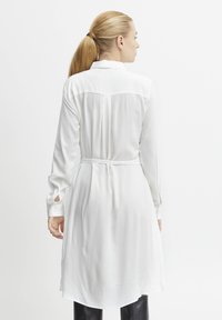 Robe blanche à manches longues avec un col, taille cintrée et un ourlet arrière légèrement allongé. Tissu lisse avec une texture minimale.