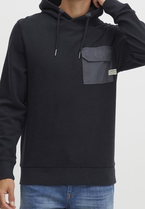 Zwarte hoodie met donkergrijze borstzak en trekkoorden, gedragen met blauwe spijkerbroek, zichtbaar tot halverwege de torso en nekgebied.