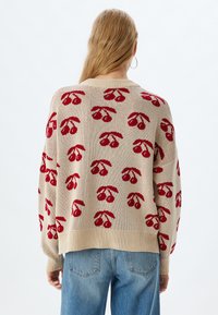 Brede gebreide trui in beige met rode kersenpatronen, ribbelmanchetten en zoom. Gepaard met lichtblauwe jeans. Zachte textuur, ontspannen pasvorm.