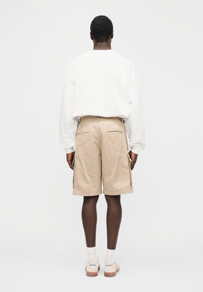 Μπεζ cargo σορτς με πλαϊνές τσέπες, σε συνδυασμό με λευκό φούτερ. Τα ρούχα έχουν άνετη εφαρμογή και είναι κατασκευασμένα από απαλό, υφασμάτινο ύφασμα με υφή.