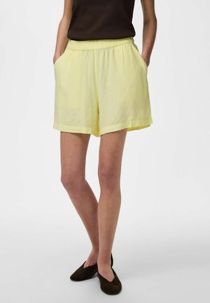 Person trägt blassgelbe Shorts mit elastischem Bund und Taschen, ein schwarzes ärmelloses Oberteil und schwarze Slipper und steht mit den Händen in den Taschen.