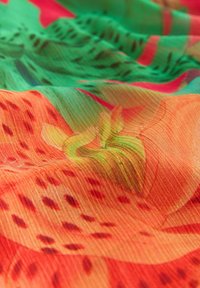 Țesătură multicolorată cu un model vibrant de flori portocalii și frunze verzi, cu textură vizibilă și pliuri sinuoase.