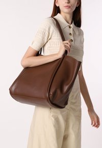 Bolso tote de cuero marrón con una textura suave, gran silueta y costuras en contraste, llevado por una persona con un atuendo de color claro.